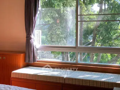 Cozy bedroom with large window and tree view, featuring built-in seating and elegant curtains.