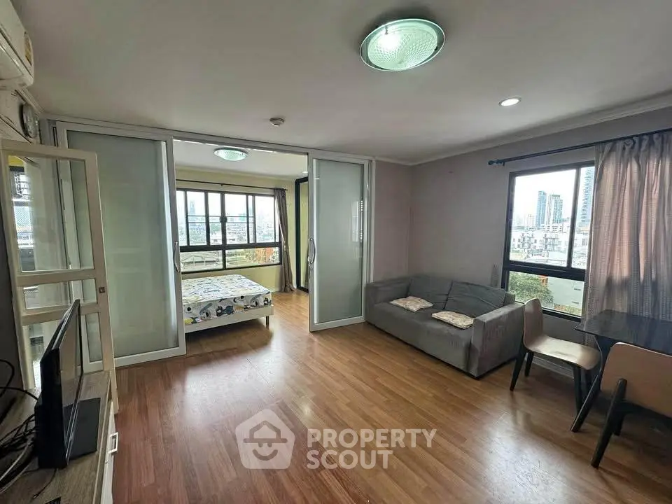 Spacious modern living room with city view, featuring wooden flooring and sliding glass doors to bedroom.