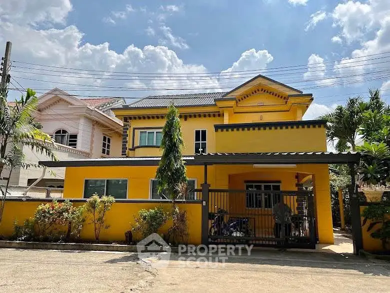 Charming yellow two-story house with gated entrance and lush greenery under a bright blue sky.