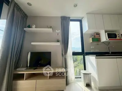 Modern living room with TV, shelves, and kitchen area featuring microwave and natural light.