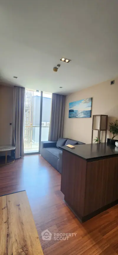 Modern living room with wooden flooring, cozy sofa, and large window leading to a balcony.