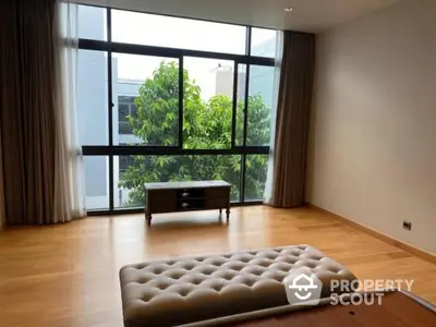 Spacious and well-lit living room with large windows offering a serene garden view, featuring polished wooden floors and elegant neutral tones.