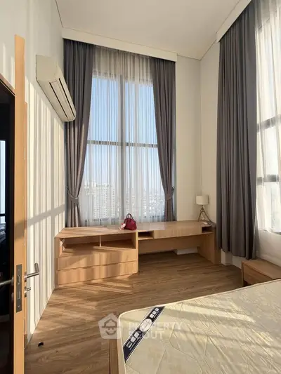 Spacious bedroom with large windows and elegant curtains, featuring a wooden desk and modern decor.