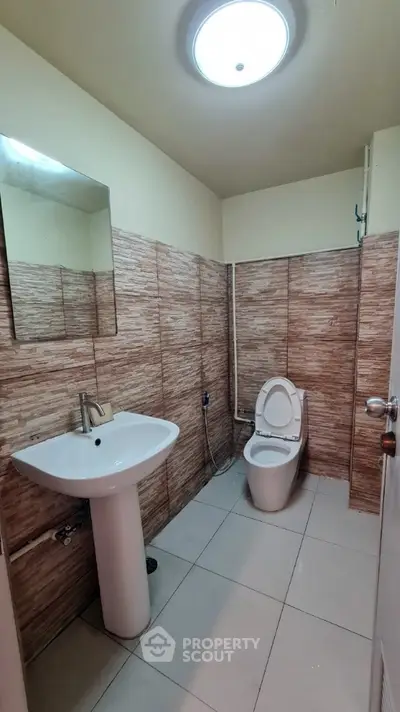Compact bathroom with modern tiled walls, pedestal sink, and well-maintained toilet under bright ceiling light.