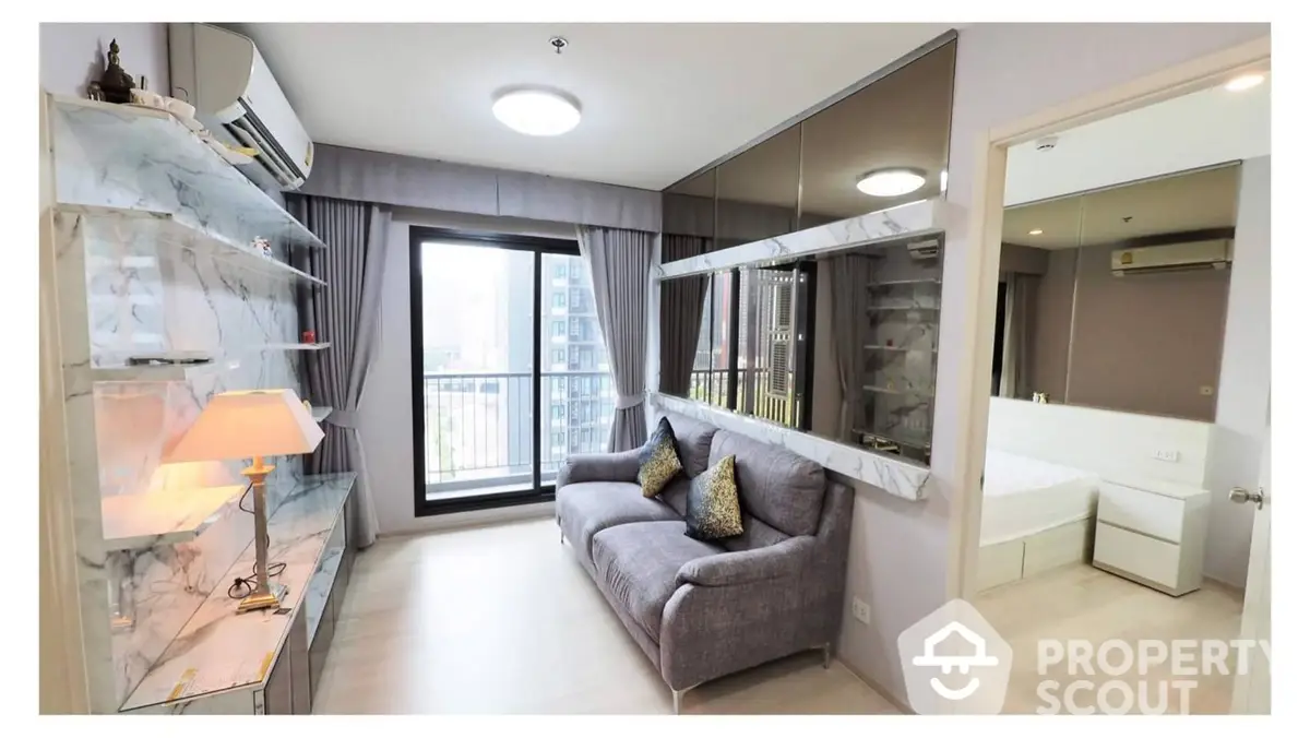 Elegant apartment interior featuring a plush grey sofa, sleek mirrored wall, and a cozy bedroom glimpse, all bathed in natural light from the balcony door.