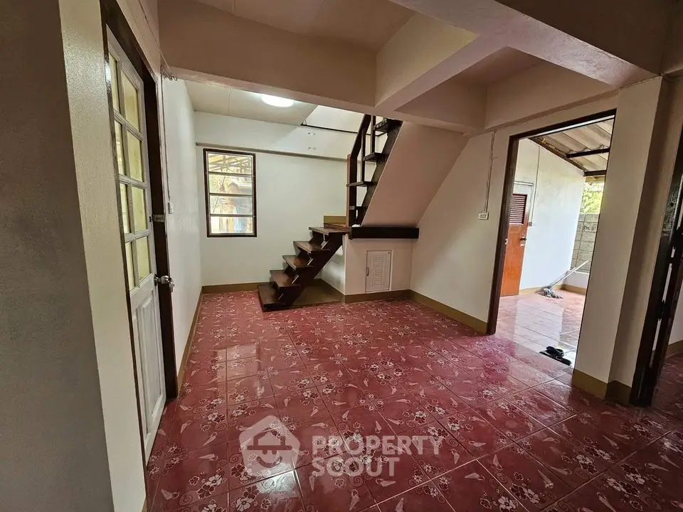 Spacious living area with striking red tiled flooring and an elegant wooden staircase leading to the upper level, surrounded by natural light from multiple windows.