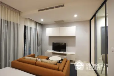 Modern living room with sleek design, featuring a plush caramel sofa, flat-screen TV, and floor-to-ceiling glass doors leading to a well-lit balcony.