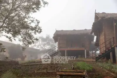 Charming rustic wooden cabins with thatched roofs in a misty countryside setting.