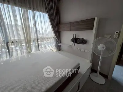 Modern bedroom with large window and city view, featuring a mounted TV bracket and standing fan.