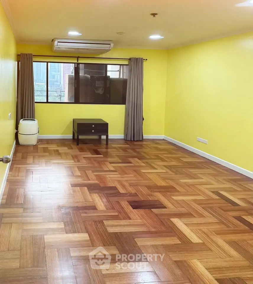 Spacious living room with parquet flooring and vibrant yellow walls, featuring a large window with city view.