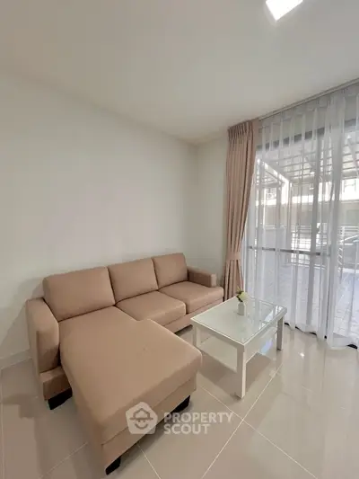 Modern living room with beige sectional sofa and glass coffee table, bright natural light.