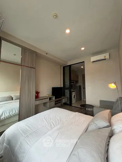 Modern bedroom with cozy bedding, mirrored wardrobe, and open view to kitchen area with appliances.