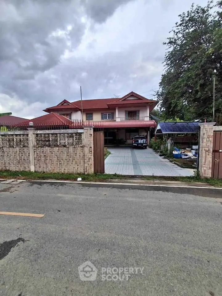 Charming two-story house with red roof and spacious driveway, surrounded by lush greenery and secure fencing.