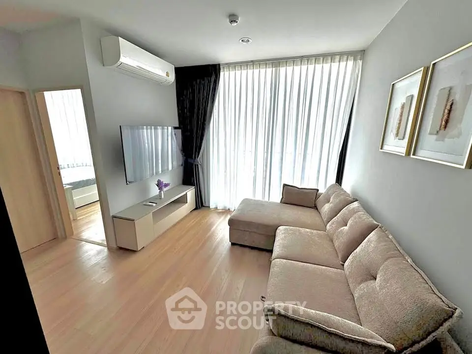Modern living room with large sofa and wall-mounted TV, featuring natural light from floor-to-ceiling windows.