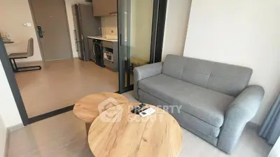 Modern living room with gray sofa and wooden tables, open kitchen layout with appliances.