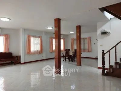 Spacious open-plan living area with dining set and staircase, featuring large windows and tiled flooring.