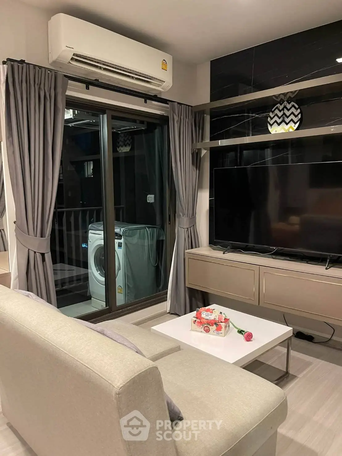 Modern living room with balcony and washing machine, featuring sleek furniture and a large TV.