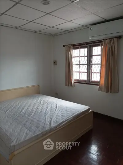 Cozy bedroom with polished wooden floors and natural light filtering through traditional window curtains, complemented by a large comfortable bed and an air conditioning unit for optimal climate control.