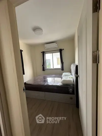 Cozy bedroom with large window and air conditioning, featuring a neatly made bed and wooden flooring.