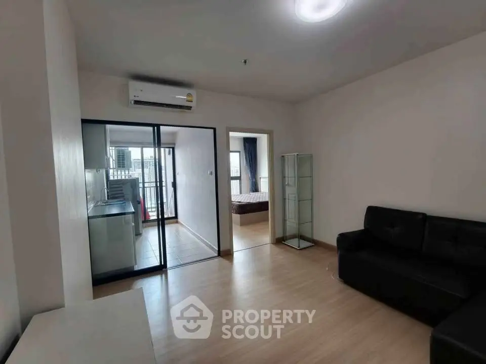 Modern apartment living room with sleek black sofa and glass cabinet, featuring a view into the bedroom and balcony.