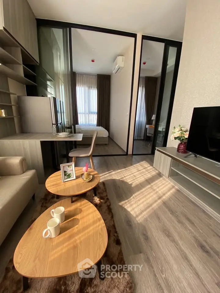 Modern living room with stylish furniture and natural light, featuring a cozy bedroom view through glass doors.