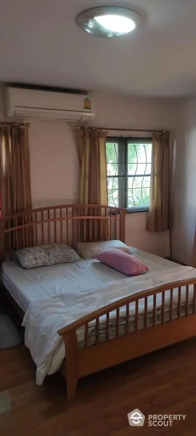 Cozy bedroom with wooden bed frame and serene greenery view through the window, complemented by warm curtains and hardwood flooring.