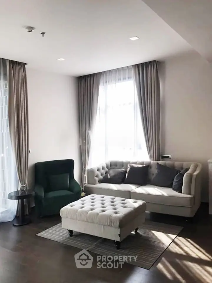 Elegant living room with plush sofa, armchair, and ottoman, featuring large windows and stylish curtains.