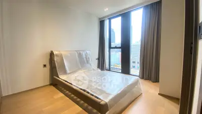 Modern bedroom with large windows and city view, featuring a new bed wrapped in protective plastic.