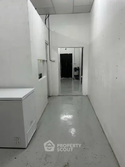 Industrial hallway with white walls and a freezer, leading to a storage area.