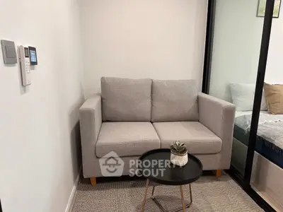 Cozy living area with modern gray sofa and small round table, adjacent to a bedroom separated by glass partition.