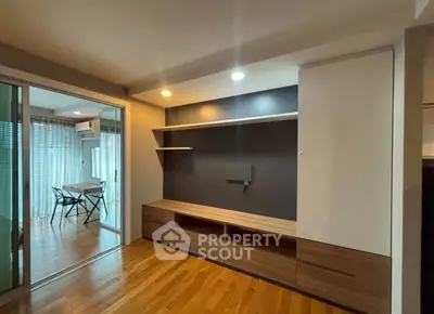 Modern living room with wooden flooring and built-in shelving, adjacent to a glass-enclosed dining area.