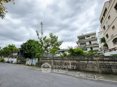 Charming residential street view with lush greenery and multi-story buildings under a cloudy sky.