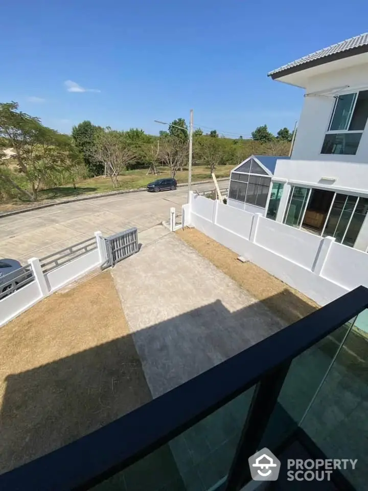 Spacious balcony with a serene view overlooking a lush green landscape and a well-paved driveway, part of a modern white residential building.