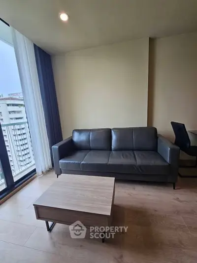 Modern living room with sleek black sofa and wooden coffee table, featuring large windows for natural light.