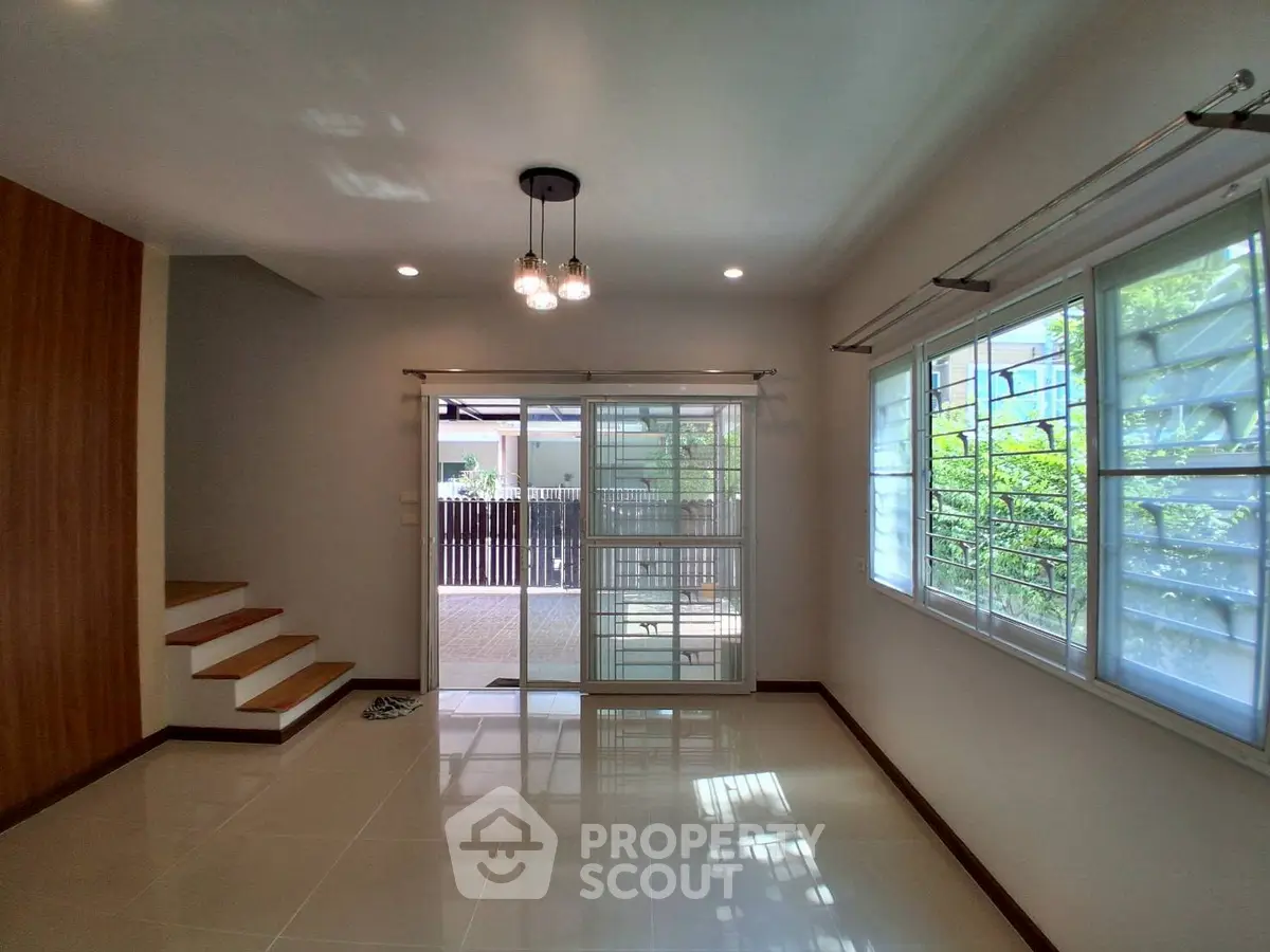 Bright living room with modern lighting and large windows overlooking a garden, featuring sleek tiled flooring.