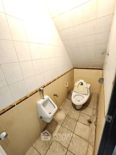 Compact bathroom with sloped ceiling and tiled walls, featuring a toilet and urinal.