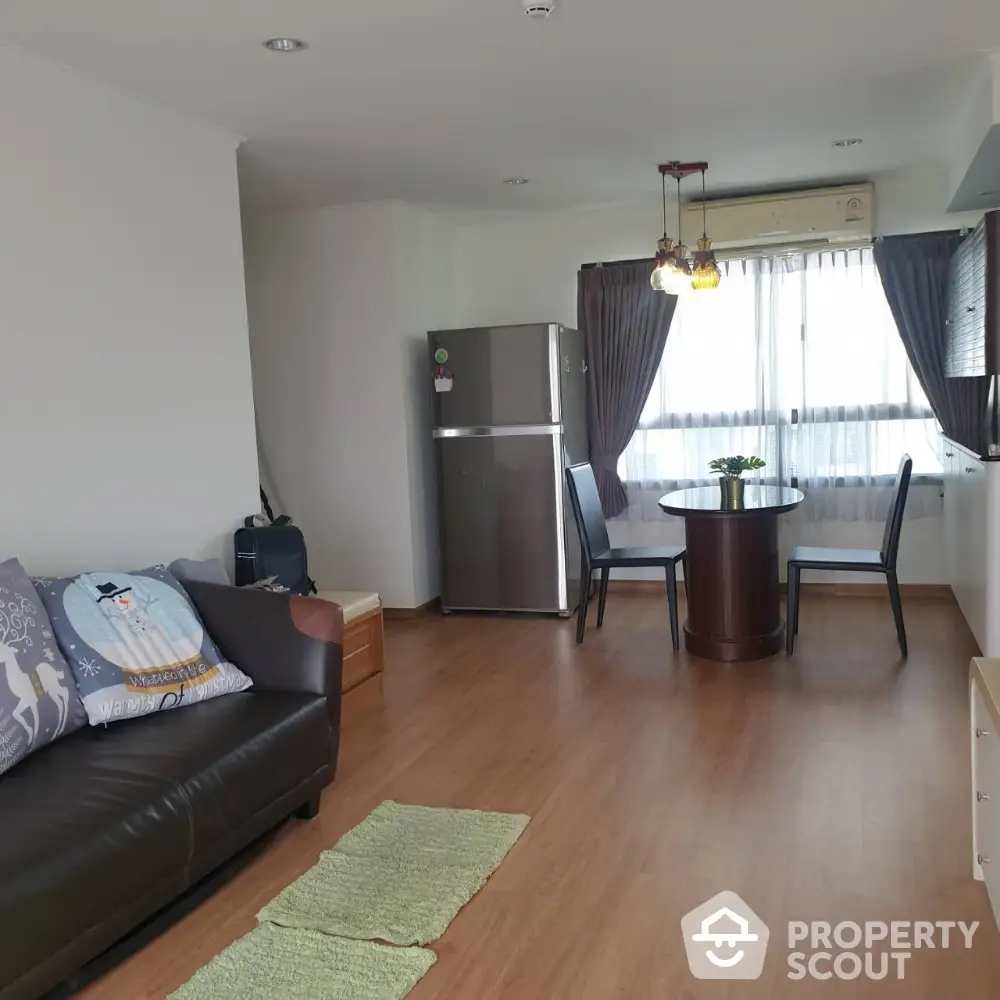 Cozy living room with dining area and modern fridge, wooden flooring, and natural light.