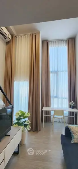 Modern living room with high ceilings and large windows, featuring elegant curtains and a cozy workspace.