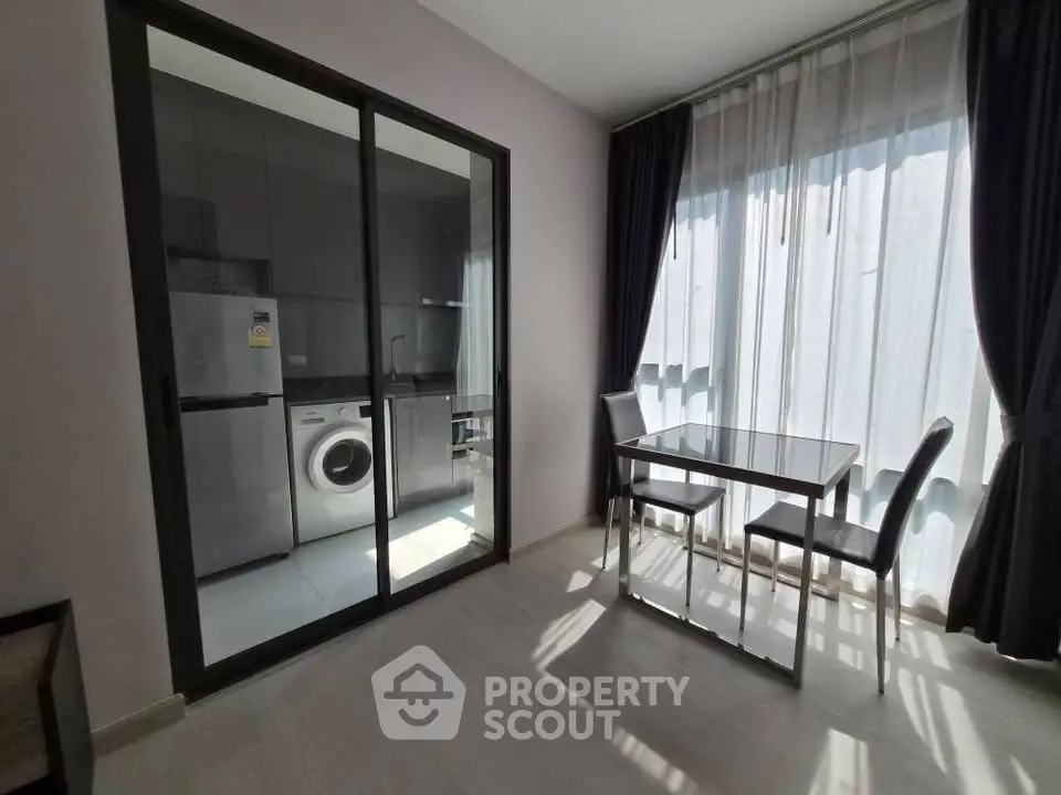 Modern apartment dining area with glass table and view into sleek kitchen with washing machine.