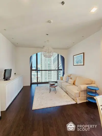 Spacious living room with hardwood floors, large windows offering city views, and elegant chandelier lighting.