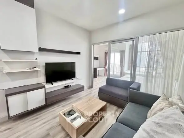 Modern living room with sleek furniture and large TV, open layout leading to a bright bedroom.