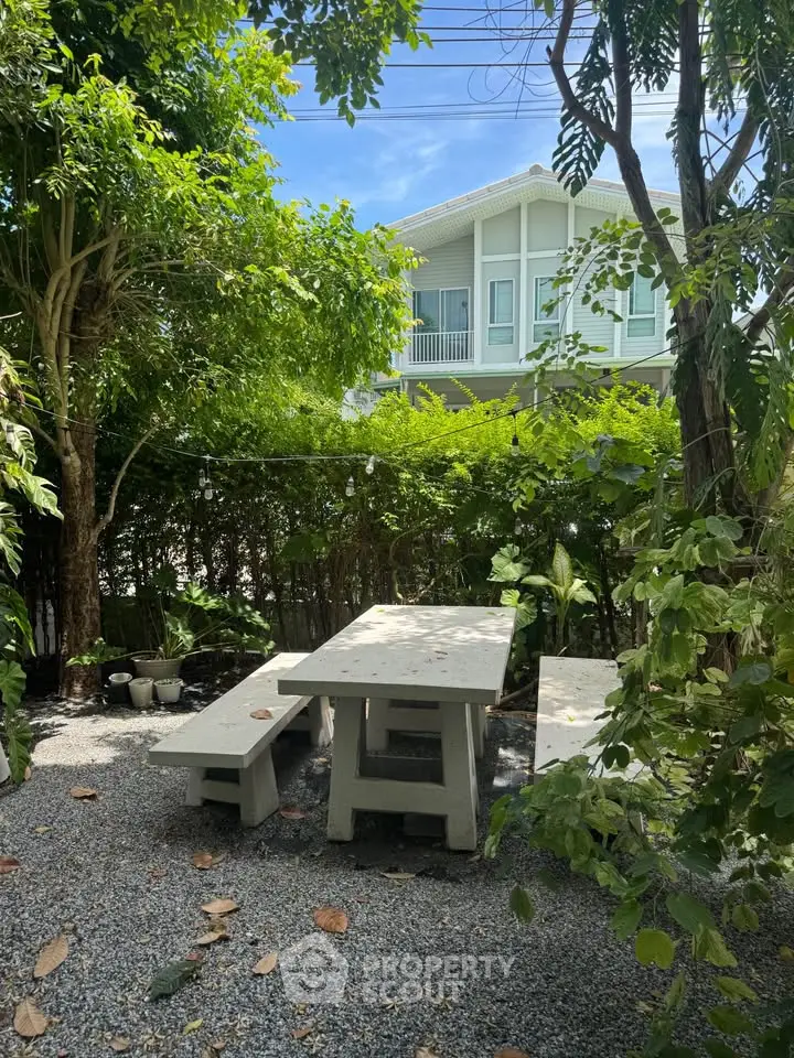 Charming garden area with rustic seating, lush greenery, and a view of a modern house.