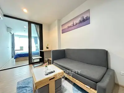 Modern living room with sleek grey sofa and wooden accents, leading to a cozy bedroom.