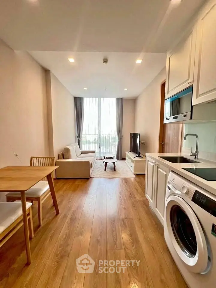 Modern living room with open kitchen and washing machine, featuring wooden flooring and natural light.