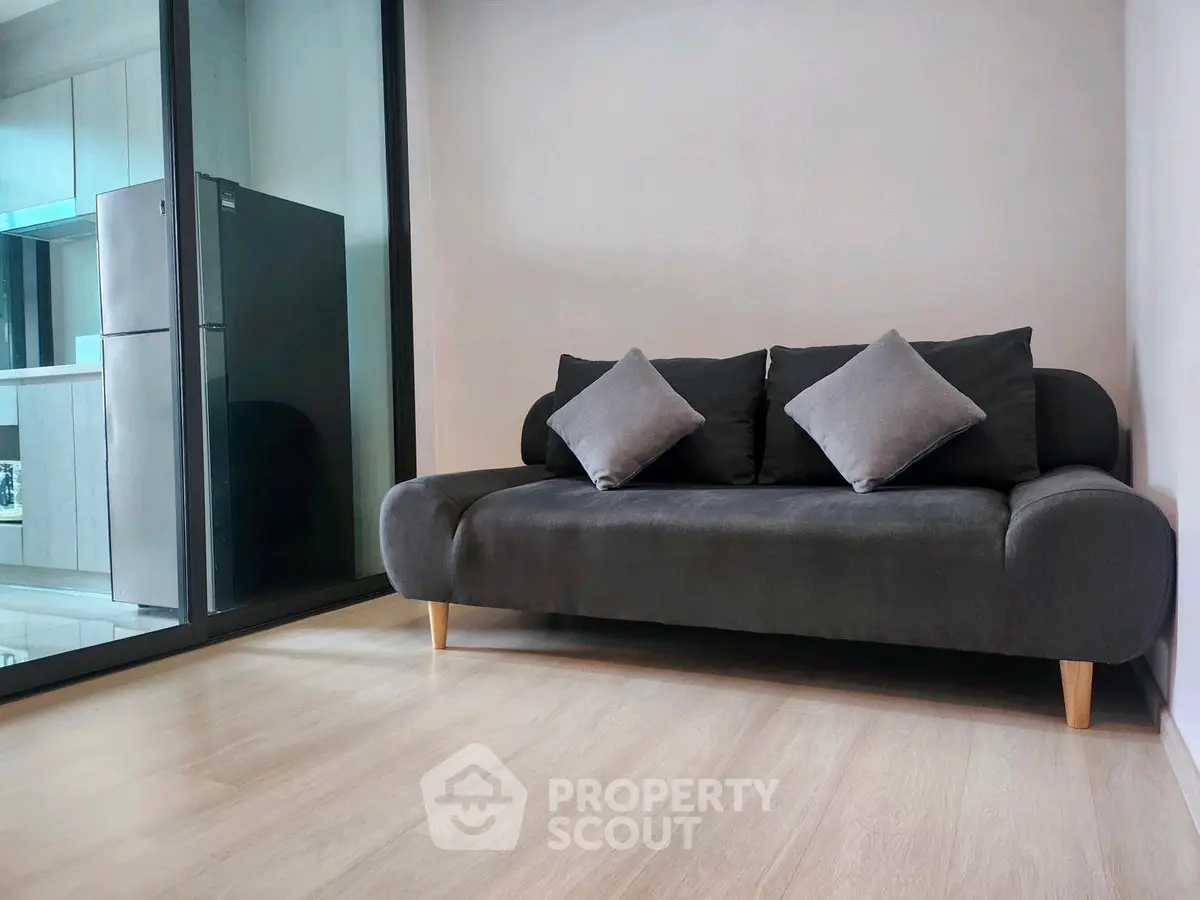 Modern living room with sleek sofa and glass partition leading to kitchen area.