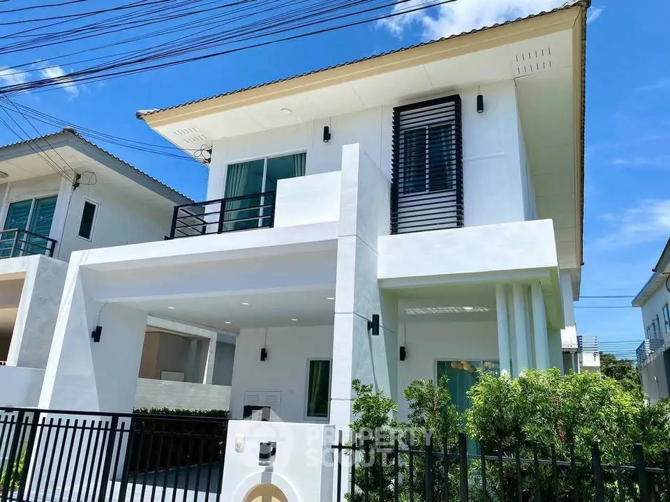 Modern two-story white house with balcony and lush greenery under a clear blue sky.