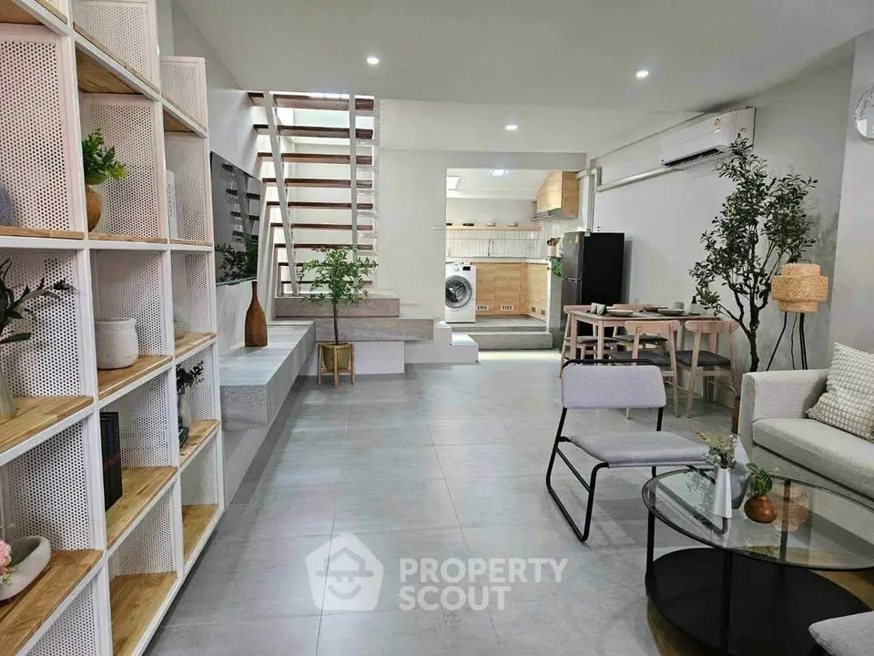 Modern open-plan living room with stylish decor and visible kitchen area, featuring sleek furniture and natural light.
