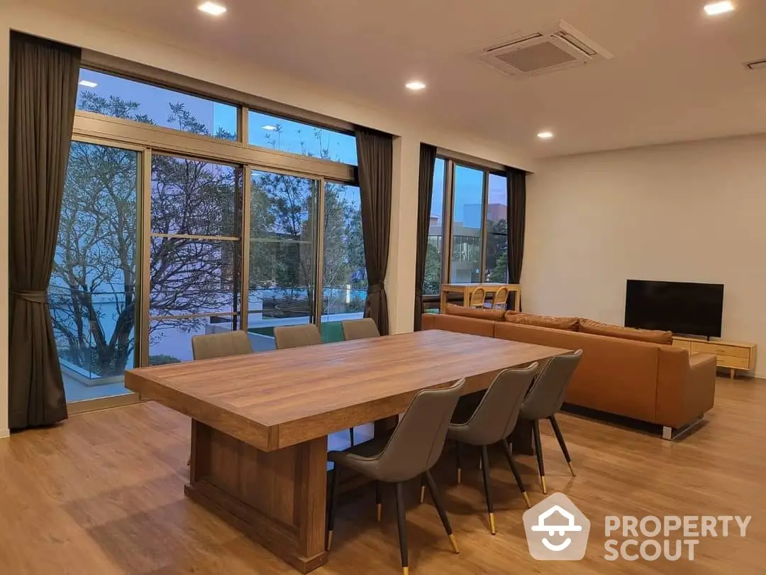 Spacious living room with large windows offering ample natural light and a serene view, complemented by a modern dining set and cozy seating area.