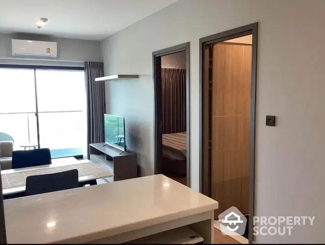 Modern apartment interior with an open-plan living space featuring a sleek white table, cozy sofa, and a flat-screen TV, bathed in natural light from large windows.