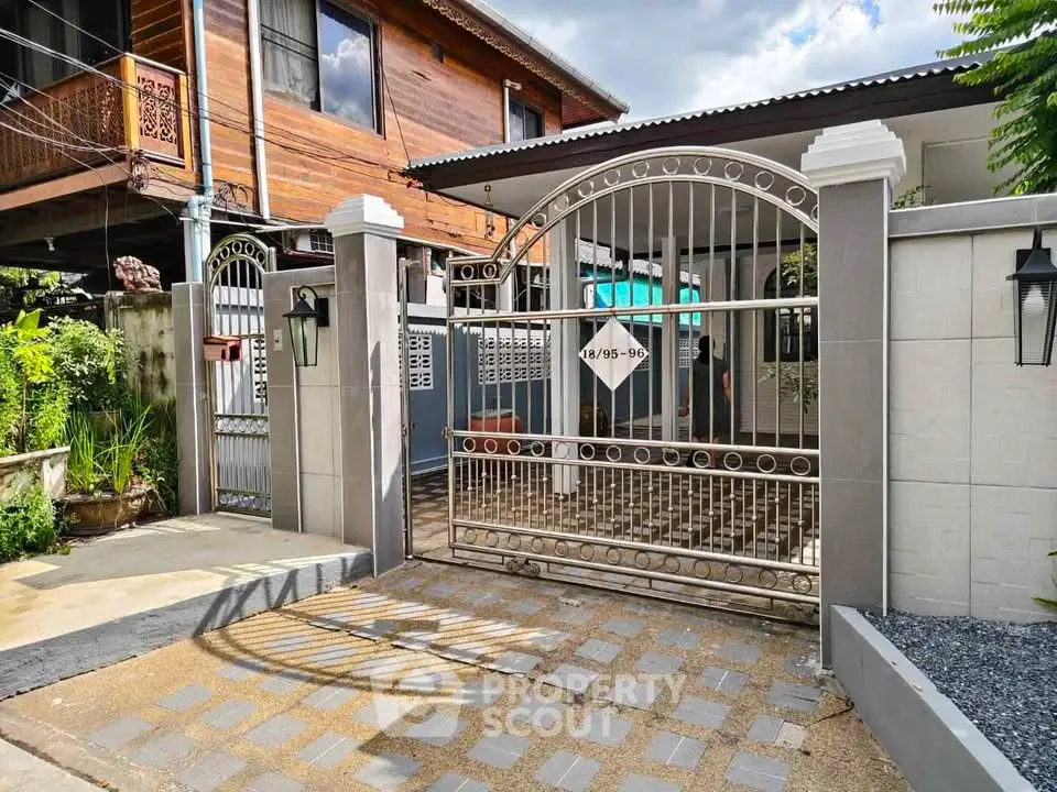 Charming gated entrance of a traditional wooden house with modern metal gate and lush greenery.